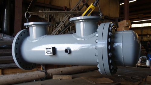 The industrial heat exchanger, featuring a cylindrical grey body and multiple flanges, showcases an efficient tube bundle system. Placed horizontally in a busy workshop, it stands among scattered tools and equipment in the background.